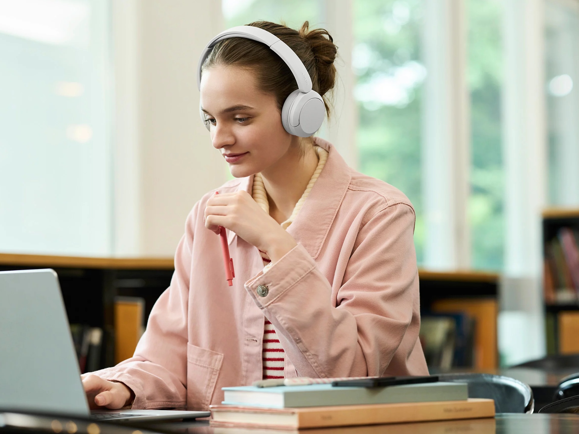 Mladá žena v růžové mikině používá bílé bezdrátové sluchátka Sony a pracuje u notebooku v kanceláři.