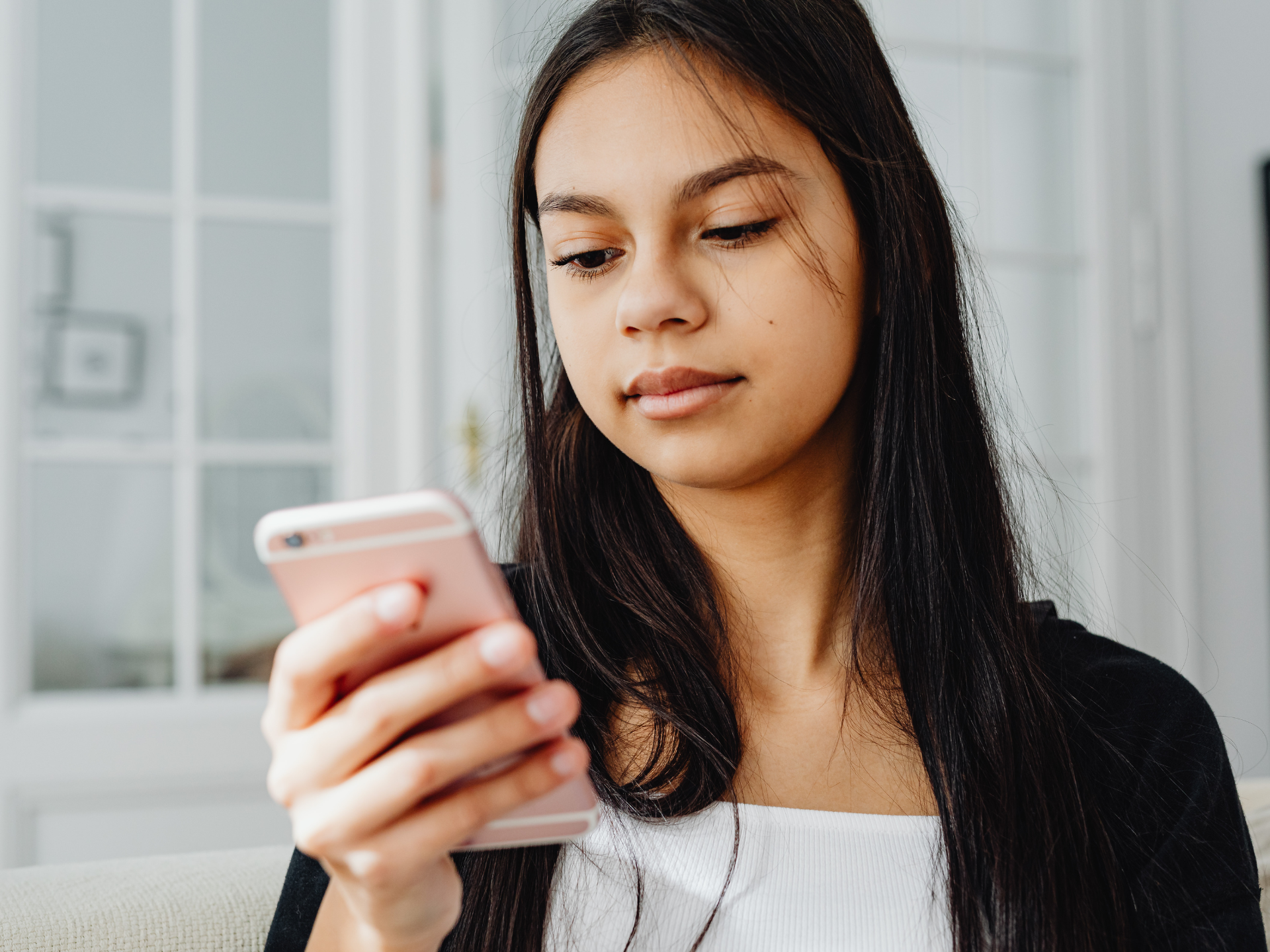 Dívka s dlouhými vlasy drží růžový smartphone, dívá se dolů, v interiéru u okna.