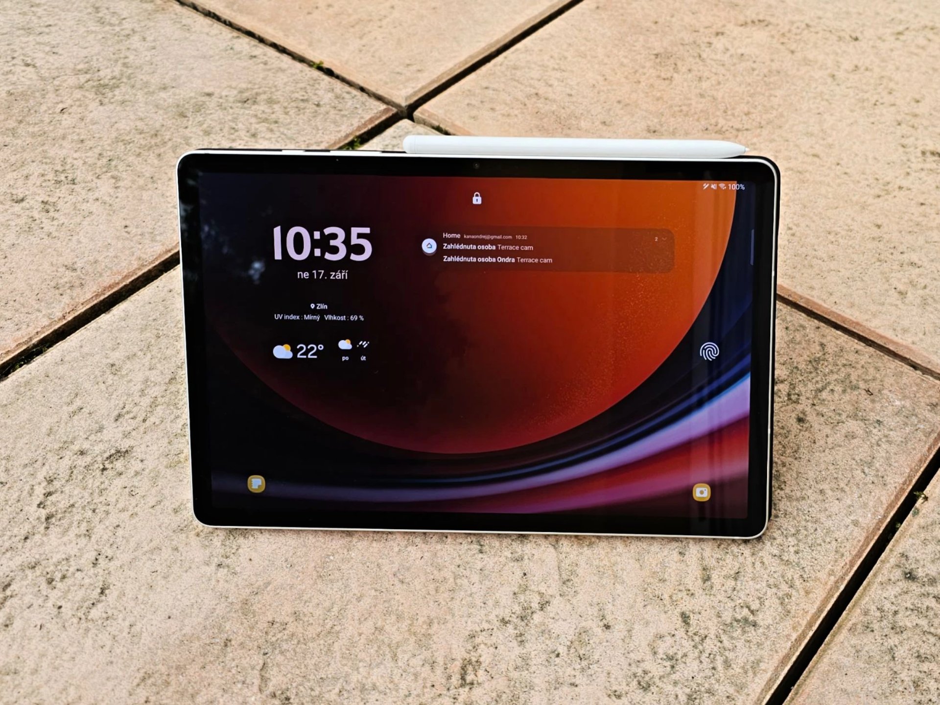 Tablet s domovskou obrazovkou na kamenné dlažbě, zobrazuje čas, počasí a připojení Wi‑Fi.