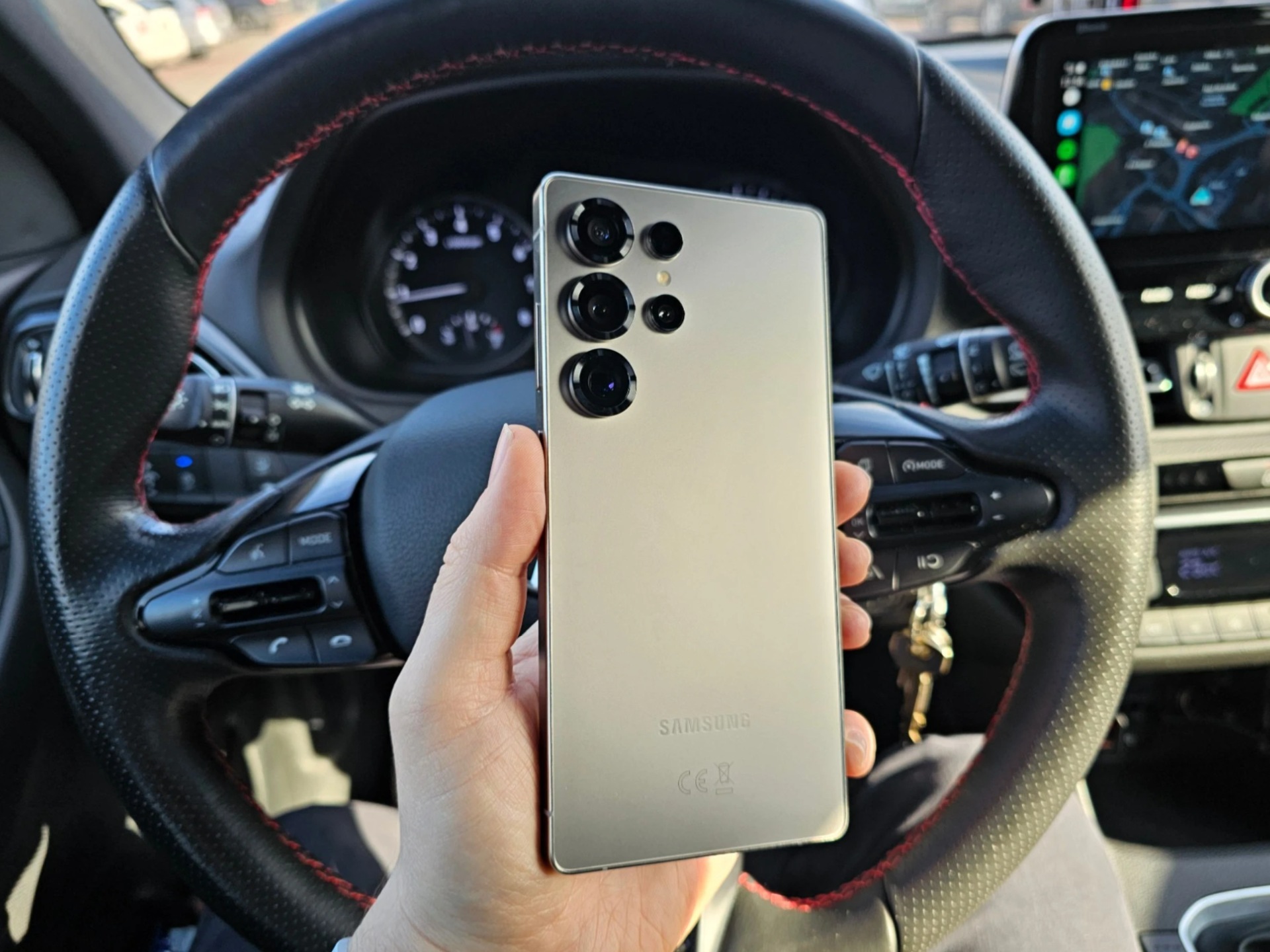 Ruka drží zadní stranu smartphonu Samsung s trojitým fotoaparátem před volantem auta.