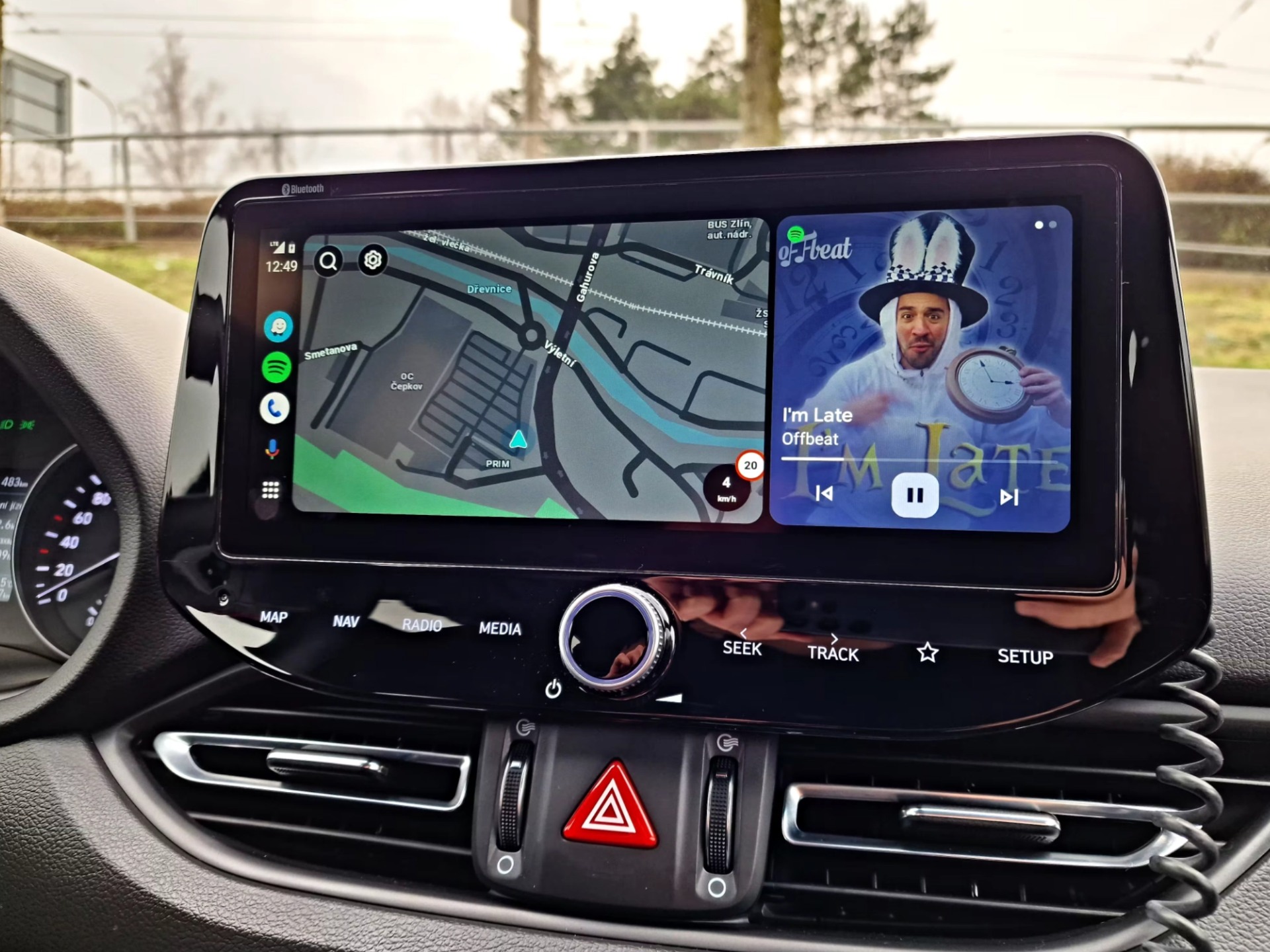 Palubní multimediální displej auta zobrazuje Android Auto a mapu navigace a hudební přehrávání, řidič drží ruku u obrazovky.