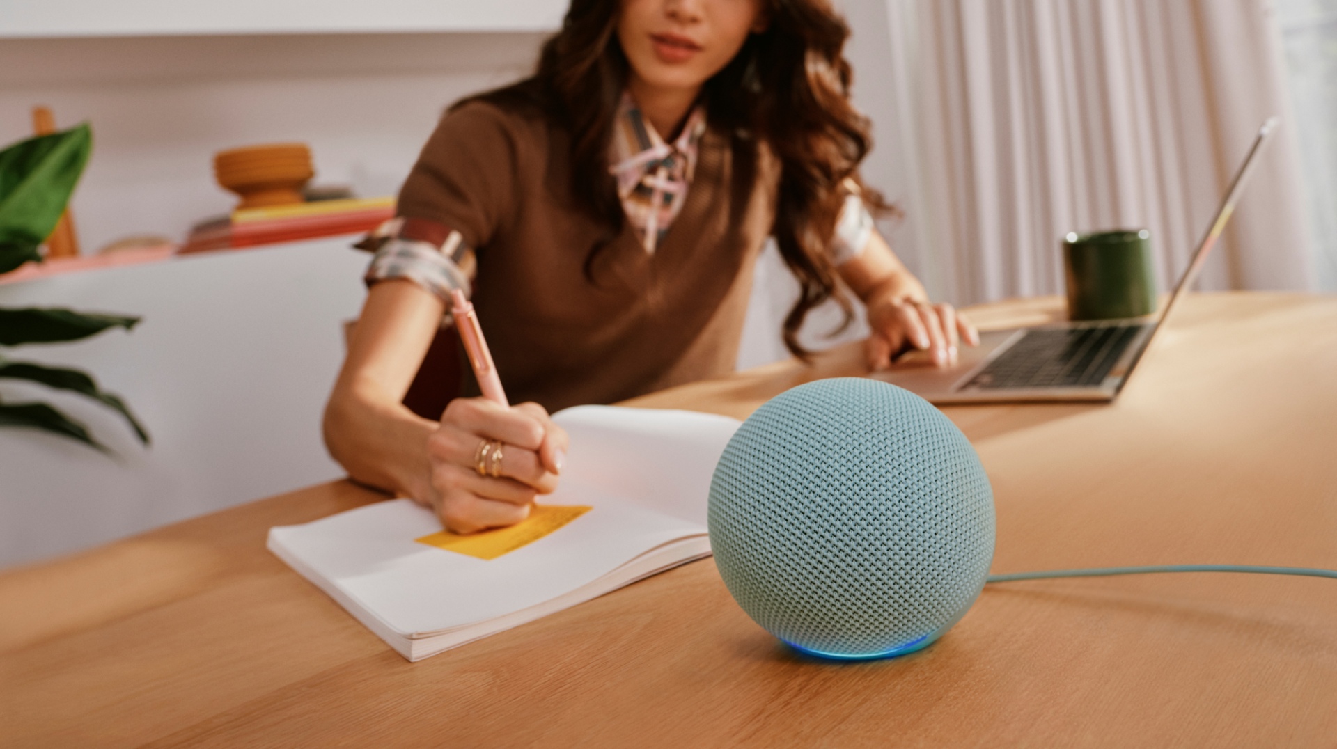 Žena sedí u stolu, píše do sešitu a vedle ní leží chytrý reproduktor Echo Dot od amazonu