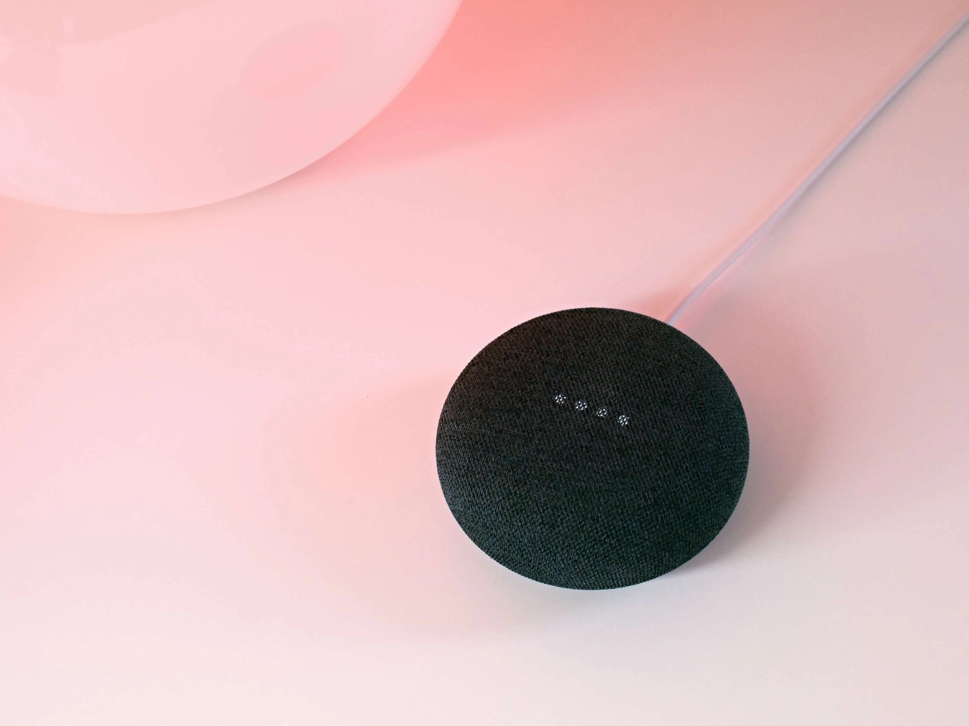 Černý kruhový reproduktor Google Nest na světle růžovém pozadí, viditelný jen horní okraj a část stínu.